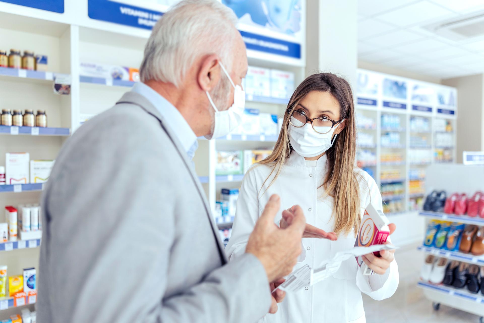 Ajuda de um farmacêutico. Uma farmacêutica com óculos possui caixas de remédios e instruções de uso. Ela olha para o homem que está explicando outra coisa. Ambos usam máscaras de proteção.